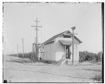 An exterior view of Exeter Substation #1 [showing distribution lines and an orchard in the background
