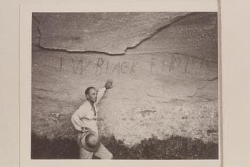 J. W. Black name with date Feb 2, 1894 written with charcoal in a cave on the right bank above the Escalante River.  Harry Aleson stands at the wall