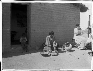 Family outside adobe structure. Woman with metate
