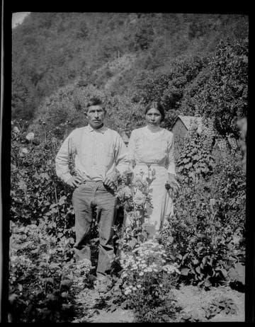 An unidentified Native American man and woman standing in a garden, with house in background