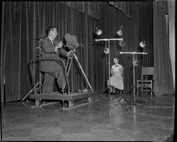 Cinematographer with 16mm movie camera on dolly filming lady seated on stage of Edison auditorium.  She's holding a card with #23 on it