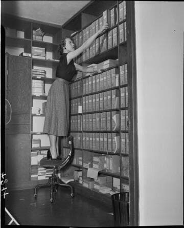 Lady standing on a swivel chair on hard floor to reach law books on top shelf of library