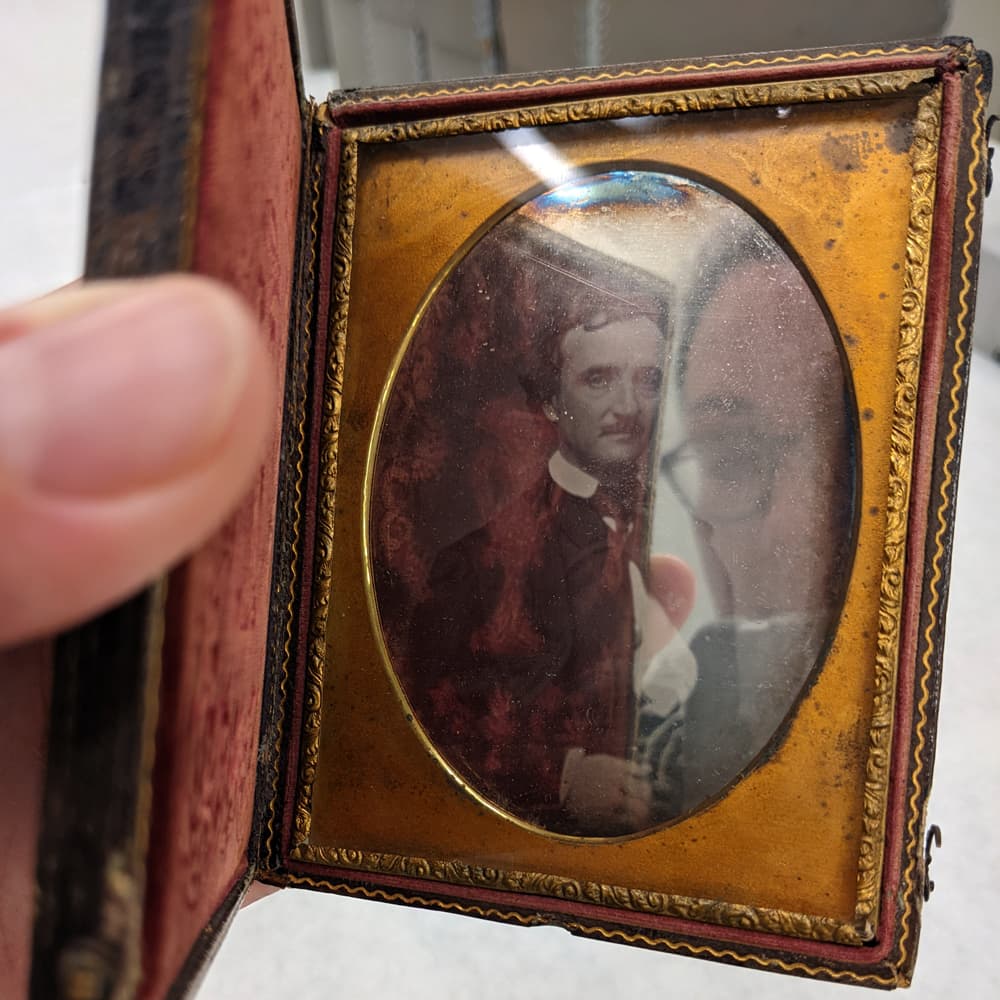 A person holds a small hinged-case with gold and velvet accents around a glass oval. Behind the glass is a formal portrait of a man; the viewer’s reflection is visible in the glass.